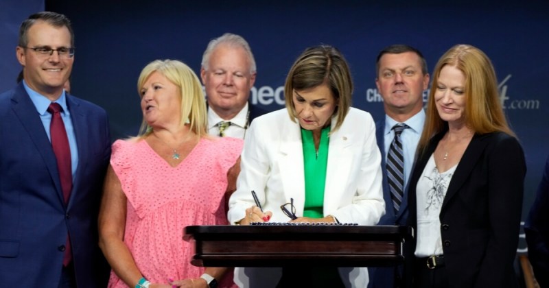 Photo is of Gov. Kim Reynolds wearing a green dress with a white jacket and bending over a podium while signing the abortion ban. Behind her are the Republican legislators who helped pass the bill. Left to right: Lt. Gov. Adam Gregg, Rep. Shannon Lundgren, Rep. Dr. Steven Bradley, Sen. Jack Whitver and Sen. Amy Sinclair