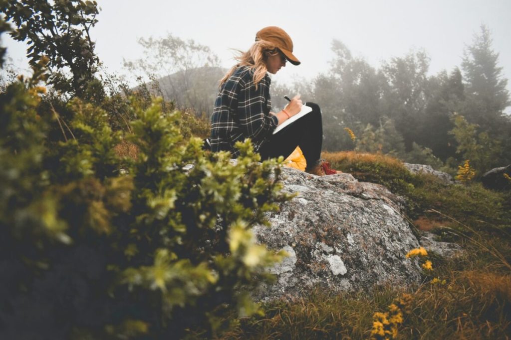 Blonde woman sitting on a rock outside writing in a notebook.