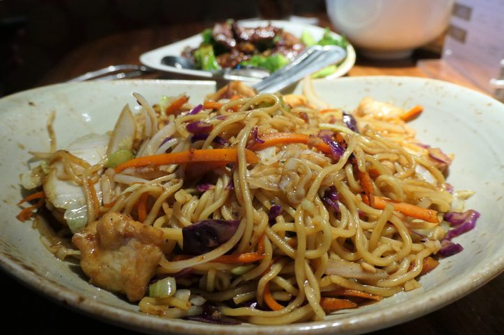 A dish of chicken lo mein has been freshly prepared for a meal. In the background is a plate of beef with broccoli.