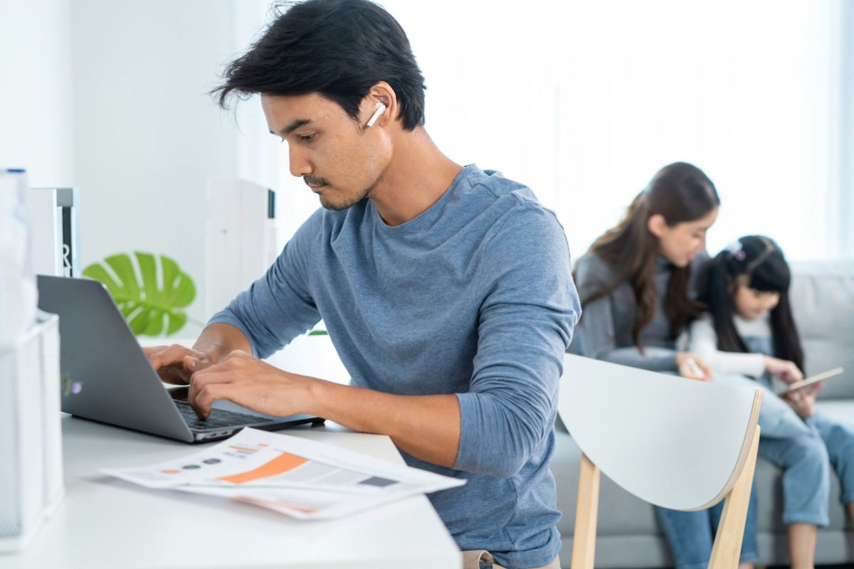 Young man in a blue long-sleeve shirt wearing headphones and working on a laptop with a woman and child in the background.